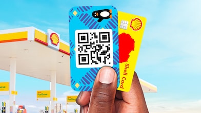 Main tenant une carte Shell et une carte Wave devant une station-service, illustrant des moyens de paiement numérique