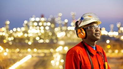 Travailleur industriel portant un équipement de sécurité devant une usine illuminée la nuit, avec casque, lunettes et protection auditive.