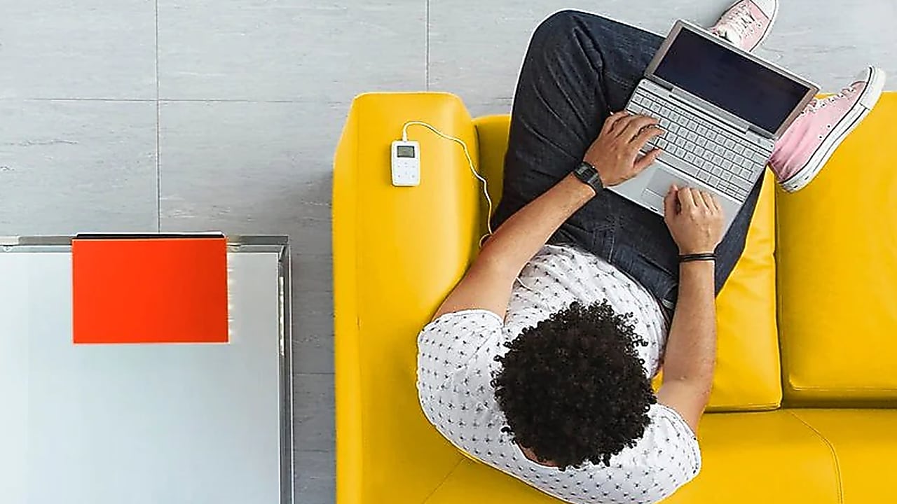 Vue plongeante d'une personne travaillant sur un ordinateur portable, assise sur un canapé jaune vif, avec un lecteur MP3 posé à côté.