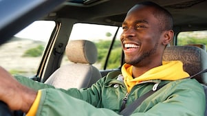 Passager souriant vêtu d'un sweat jaune et d'une veste verte assis dans une voiture pendant un trajet ensoleillé.