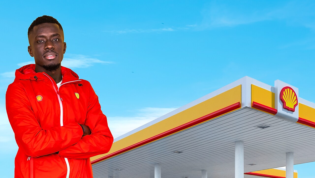 Idrissa Gana Gueye portant une veste rouge Shell devant une station-service Shell, sous un ciel bleu clair.