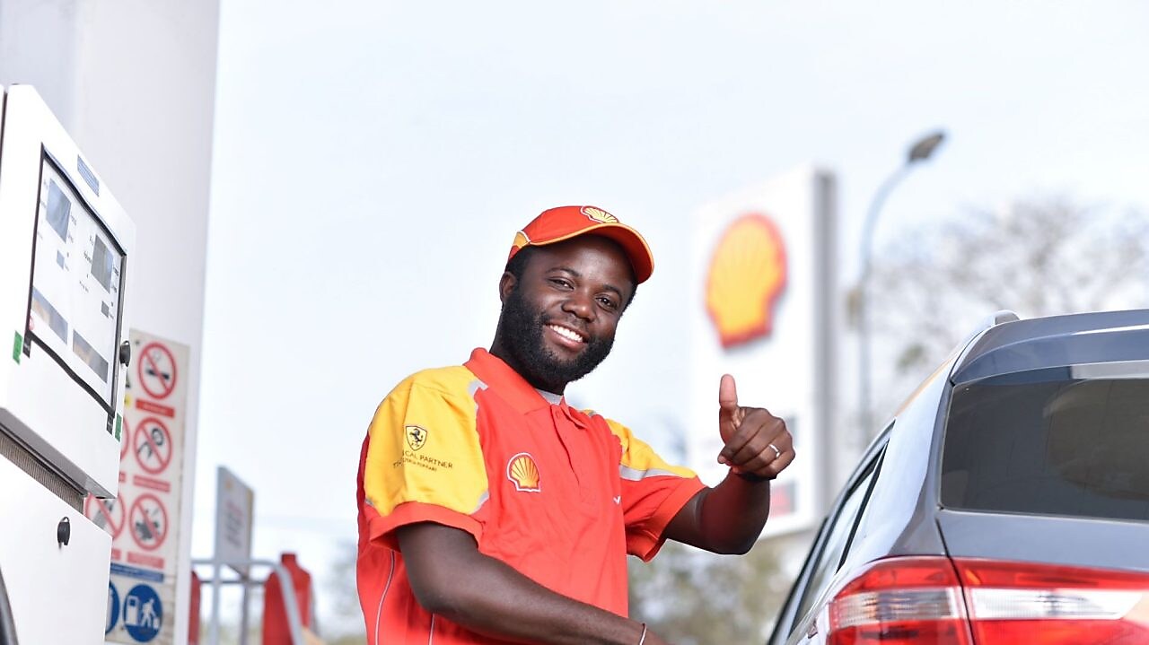 Employé souriant en uniforme Shell fait le plein d'essence à une pompe, avec le logo Shell visible en arrière-plan.