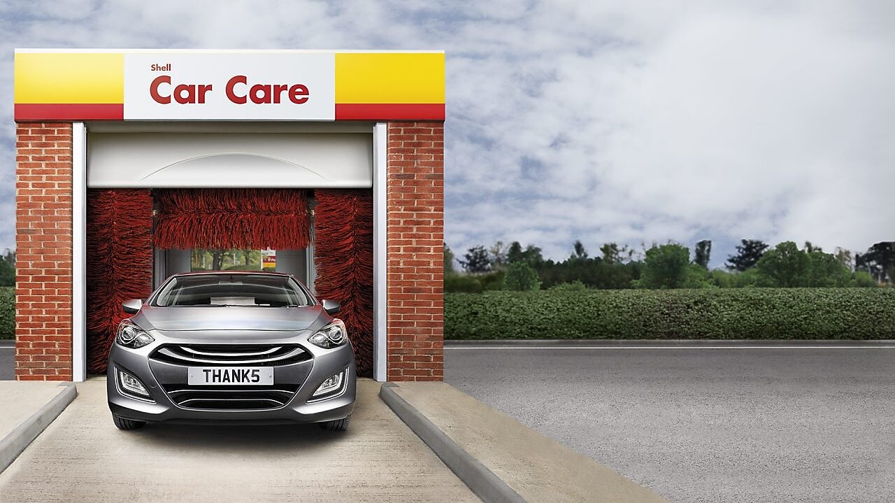 Voiture grise à l'entrée d'un tunnel de lavage Shell Car Care avec brosses rouges et plaque d'immatriculation "THANKS"