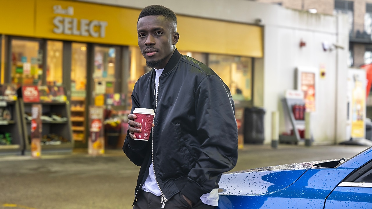 Idrissa Gana Gueye en blouson noir tenant un café devant une boutique Select, appuyé contre une voiture bleue