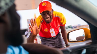 Employé Shell en uniforme rouge et jaune saluant un client à travers la fenêtre d'une voiture dans une station-service
