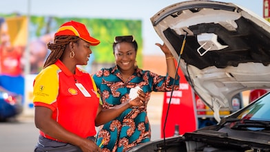 Employée de station-service Shell en uniforme rouge et jaune vérifiant le niveau d'huile d'une voiture avec une cliente souriante