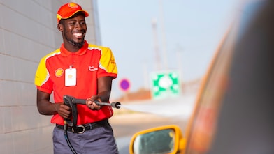 Employé souriant en uniforme Shell rouge et jaune tenant un pistolet de carburant dans une station-service