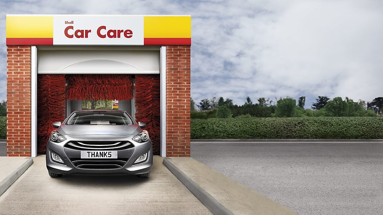 Voiture grise à l'entrée d'un tunnel de lavage Shell Car Care avec brosses rouges et plaque d'immatriculation "THANKS"