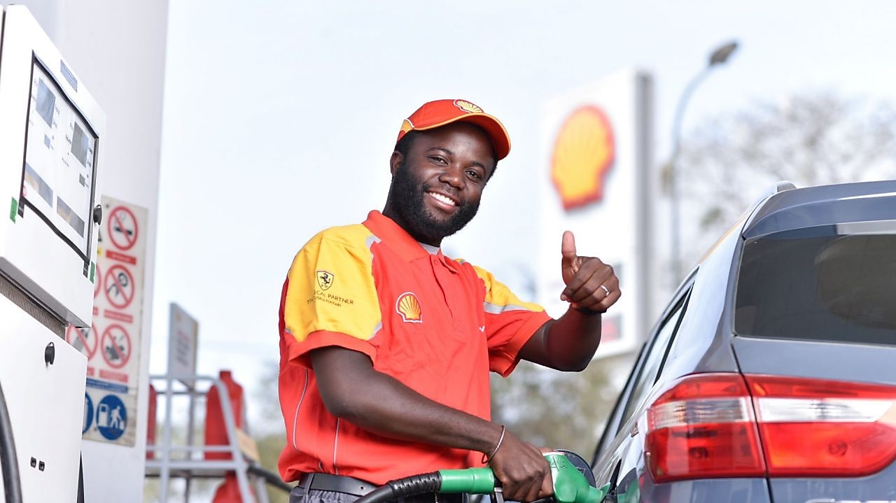 Employé souriant en uniforme Shell fait le plein d'essence à une pompe, avec le logo Shell visible en arrière-plan.