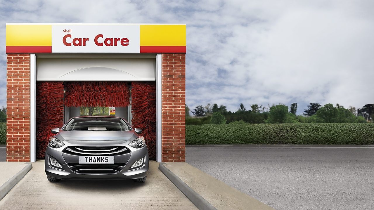 Voiture grise à l'entrée d'un tunnel de lavage Shell Car Care avec brosses rouges et plaque d'immatriculation "THANKS"