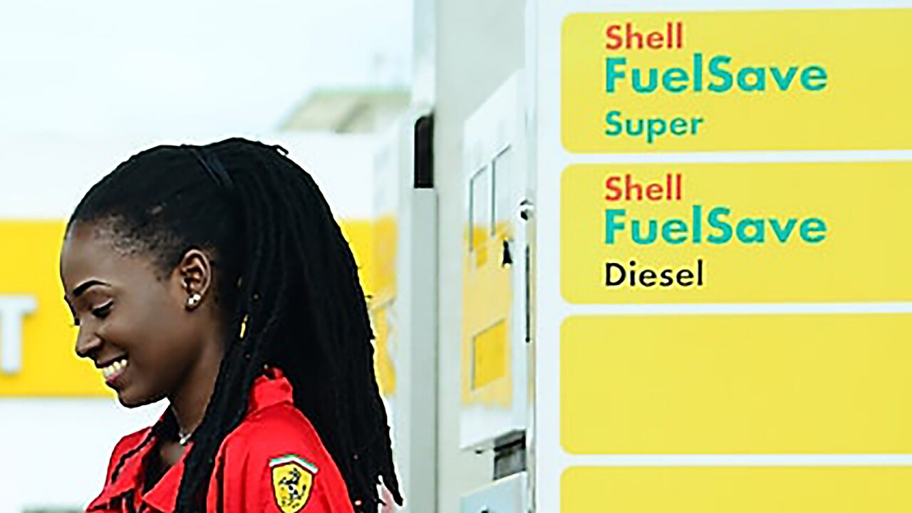 Employée en uniforme rouge Ferrari devant les pompes Shell FuelSave Super et Diesel dans une station-service
