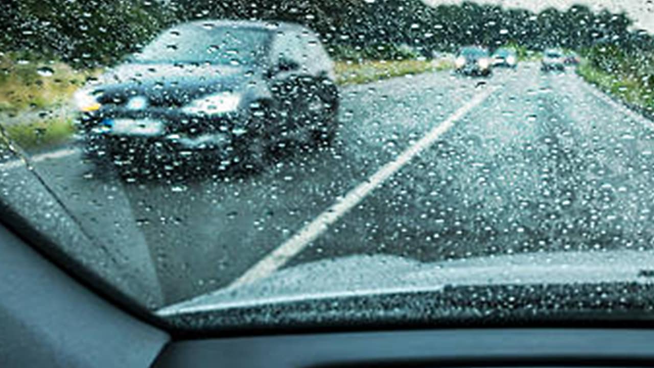 Vue à travers un pare-brise mouillé sous la pluie, avec les essuie-glaces en action et une voiture floue visible devant.