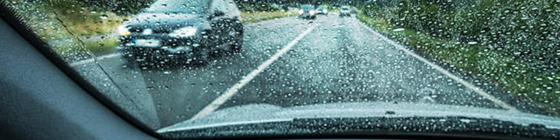 Vue à travers un pare-brise mouillé sous la pluie, avec les essuie-glaces en action et une voiture floue visible devant.