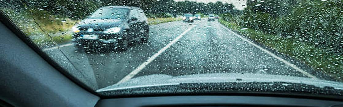 Vue à travers un pare-brise mouillé sous la pluie, avec les essuie-glaces en action et une voiture floue visible devant.
