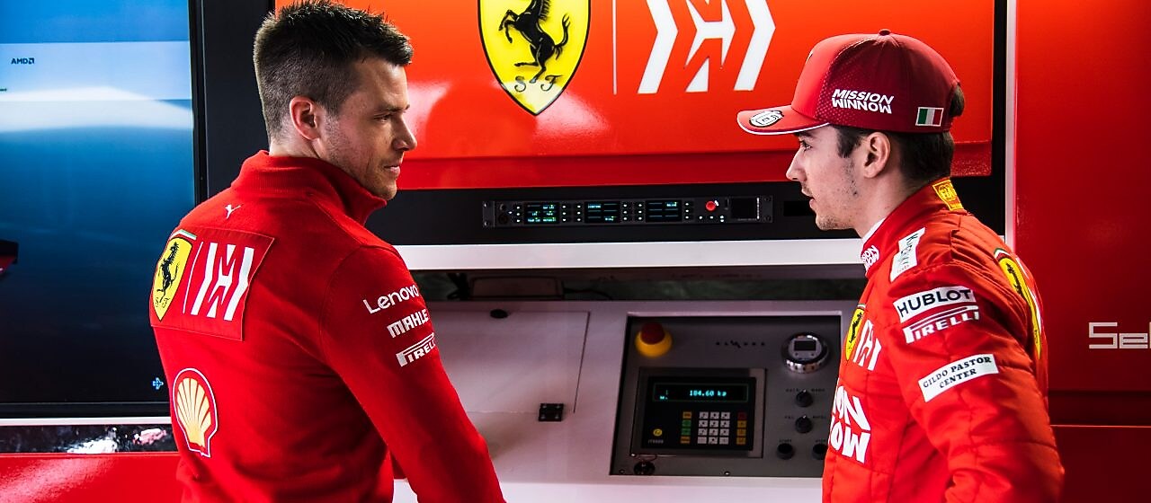 Deux membres de l'équipe Ferrari en tenue officielle discutent dans le garage de F1, avec le logo Ferrari en arrière-plan.