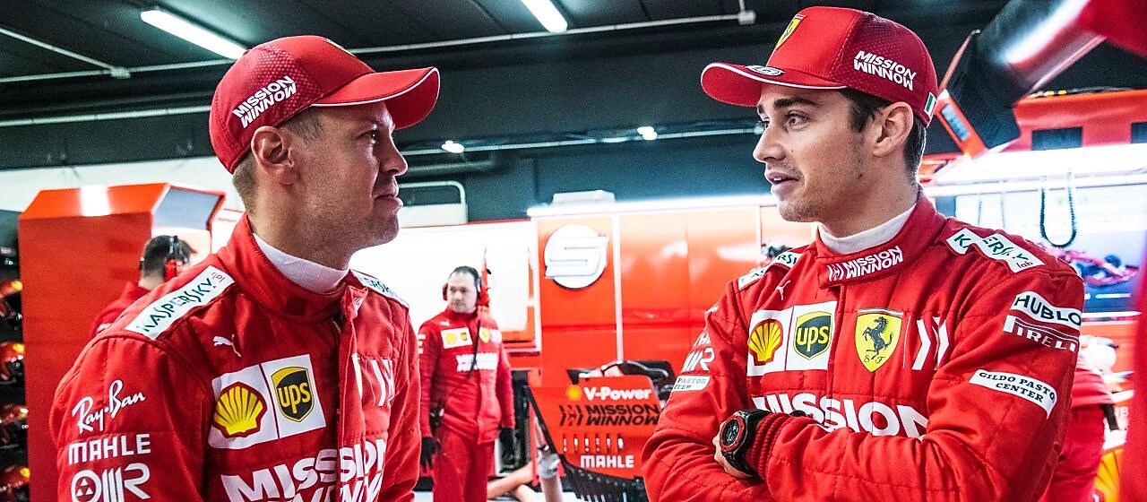Deux pilotes Ferrari en combinaison rouge et casquette discutent dans le garage de l'équipe pendant un Grand Prix de F1