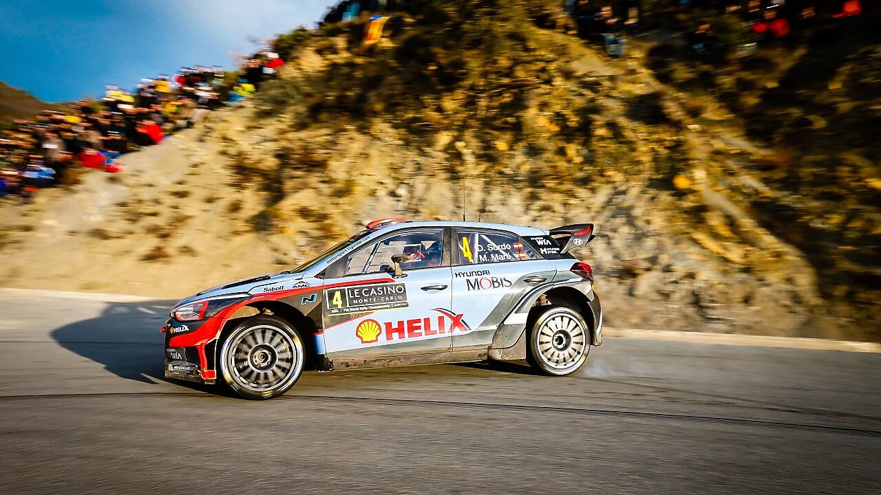Voiture de rallye Hyundai i20 WRC en action lors du Rallye Monte-Carlo, avec des spectateurs sur la falaise en arrière-plan