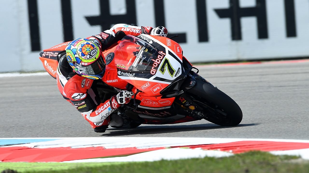 Moto Ducati rouge numéro 7 penchée dans un virage sur circuit, avec pilote portant un casque coloré