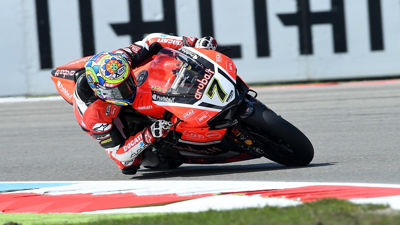 Moto Ducati rouge numéro 7 penchée dans un virage sur circuit, avec pilote portant un casque coloré