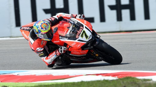 Moto Ducati rouge numéro 7 penchée dans un virage sur circuit, avec pilote portant un casque coloré