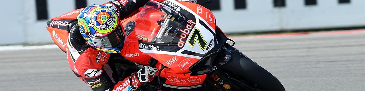 Moto Ducati rouge numéro 7 penchée dans un virage sur circuit, avec pilote portant un casque coloré