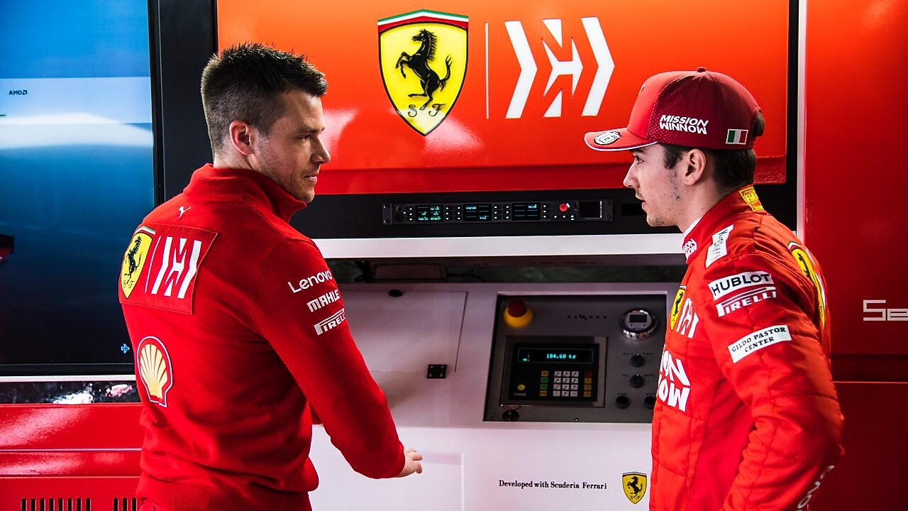 Deux membres de l'équipe Ferrari en tenue officielle discutent dans le garage de F1, avec le logo Ferrari en arrière-plan.