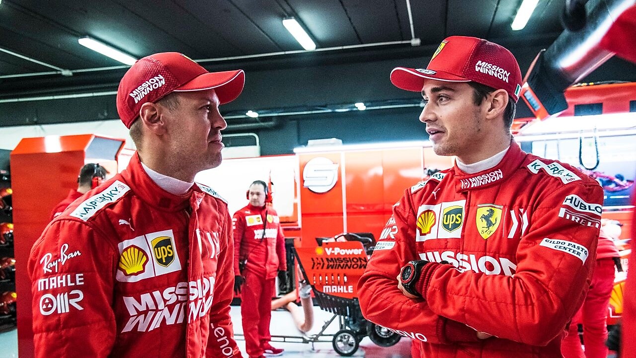 Deux pilotes Ferrari en combinaison rouge et casquette discutent dans le garage de l'équipe pendant un Grand Prix de F1