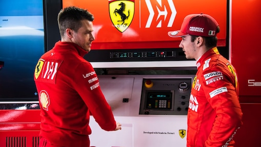 Deux membres de l'équipe Ferrari en tenue officielle discutent dans le garage de F1, avec le logo Ferrari en arrière-plan.