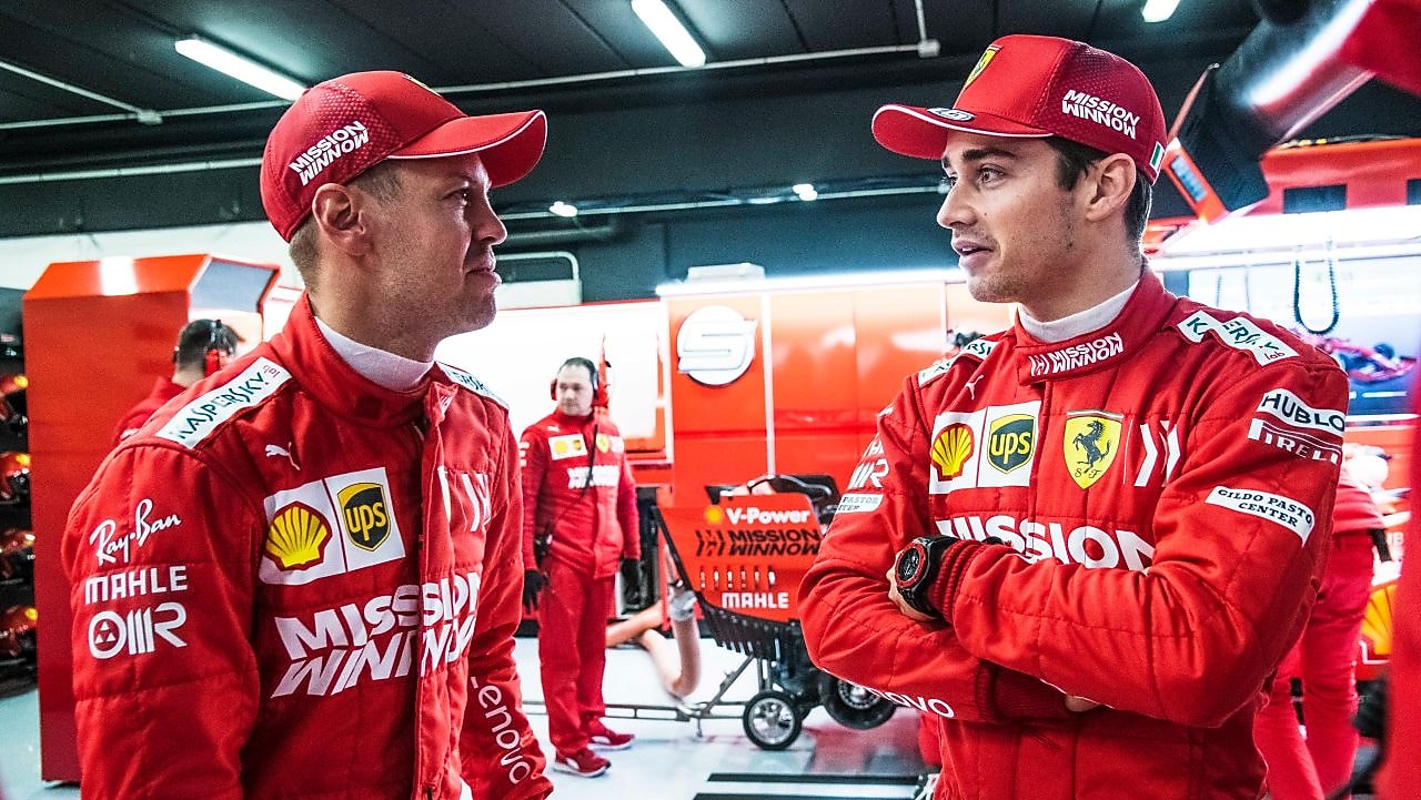 Deux pilotes Ferrari en combinaison rouge et casquette discutent dans le garage de l'équipe pendant un Grand Prix de F1