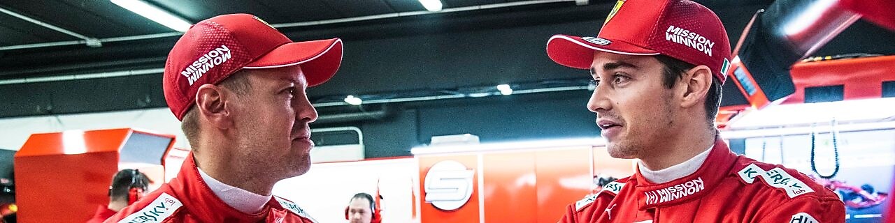 Deux pilotes Ferrari en combinaison rouge et casquette discutent dans le garage de l'équipe pendant un Grand Prix de F1