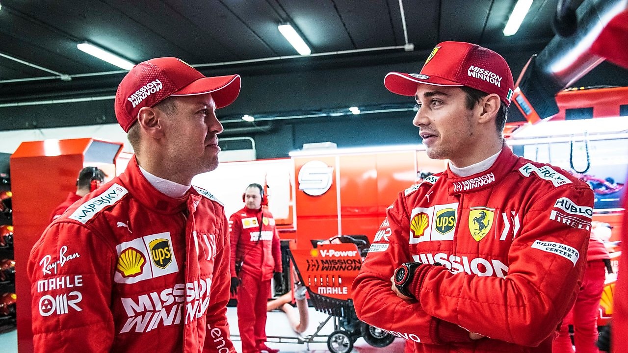 Deux pilotes Ferrari en combinaison rouge et casquette discutent dans le garage de l'équipe pendant un Grand Prix de F1