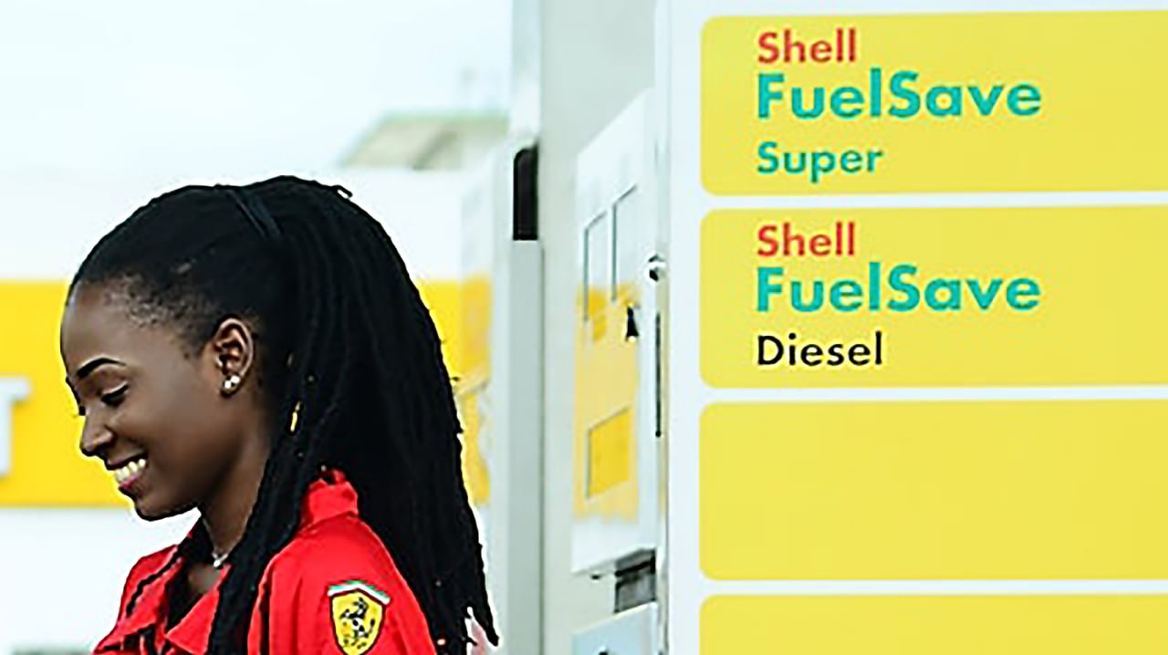 Employée en uniforme rouge Ferrari devant les pompes Shell FuelSave Super et Diesel dans une station-service