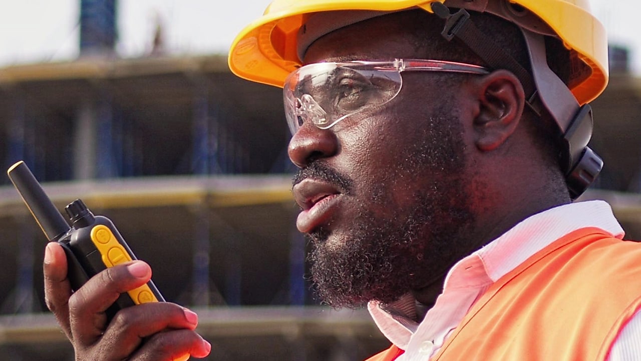 Ouvrier du bâtiment portant un casque jaune et un gilet orange, utilisant un talkie-walkie sur un chantier de construction.