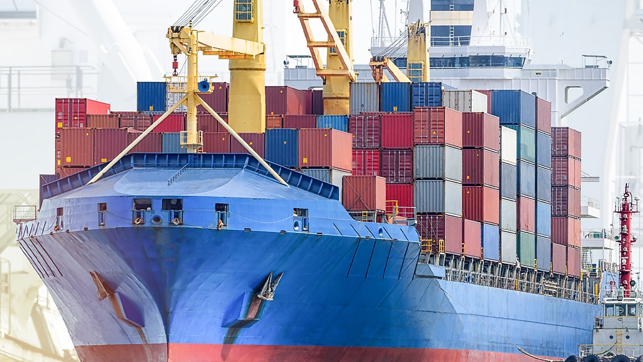 Navire porte-conteneurs bleu chargé de conteneurs colorés, avec grues de chargement jaunes dans un port commercial.