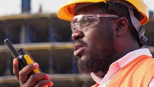 Ouvrier du bâtiment portant un casque jaune et un gilet orange, utilisant un talkie-walkie sur un chantier de construction.